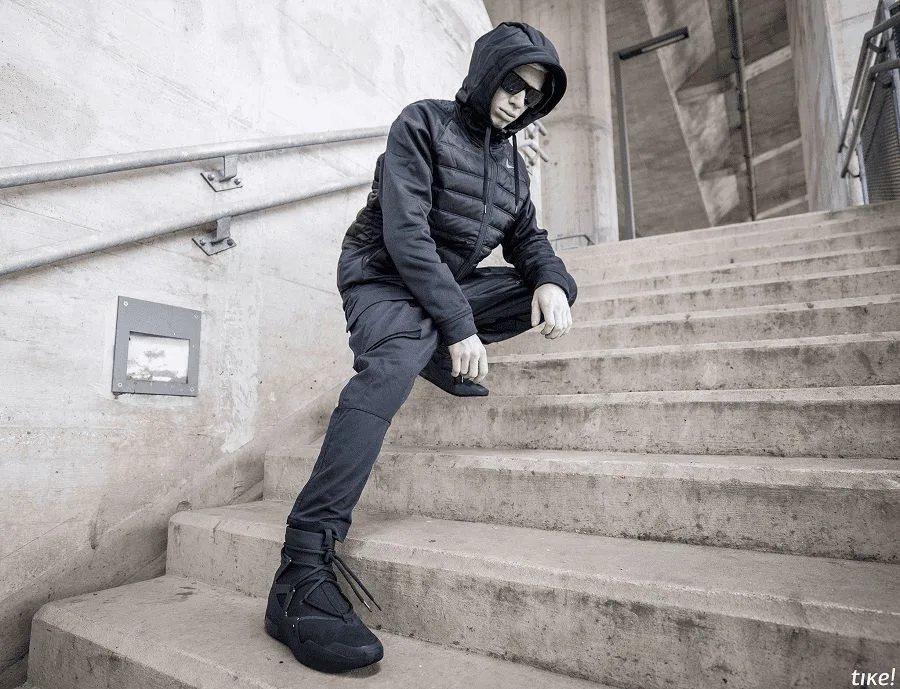 The same all-black outfit shot from below on a wide concrete staircase, one Nike Air Fear of God 1 boot planted on the step in front