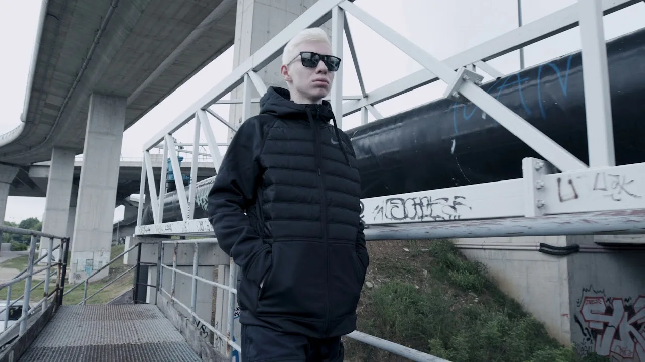 Still from the Tike lookbook film: the model in a black jacket standing on a gangway under a concrete overpass
