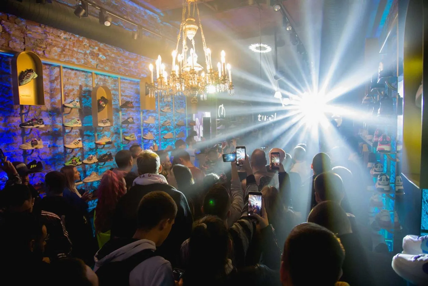 A packed room inside the Tike store under a chandelier, phones raised above the crowd, sneakers displayed in lit alcoves along the stone wall