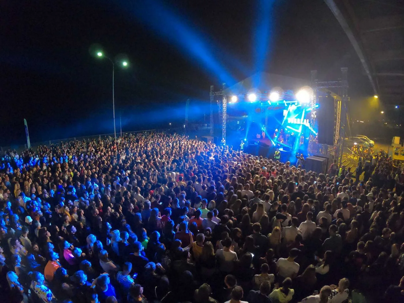 A dense night-time crowd in front of a lit stage at the Port of Belgrade, blue beams cutting over thousands of heads