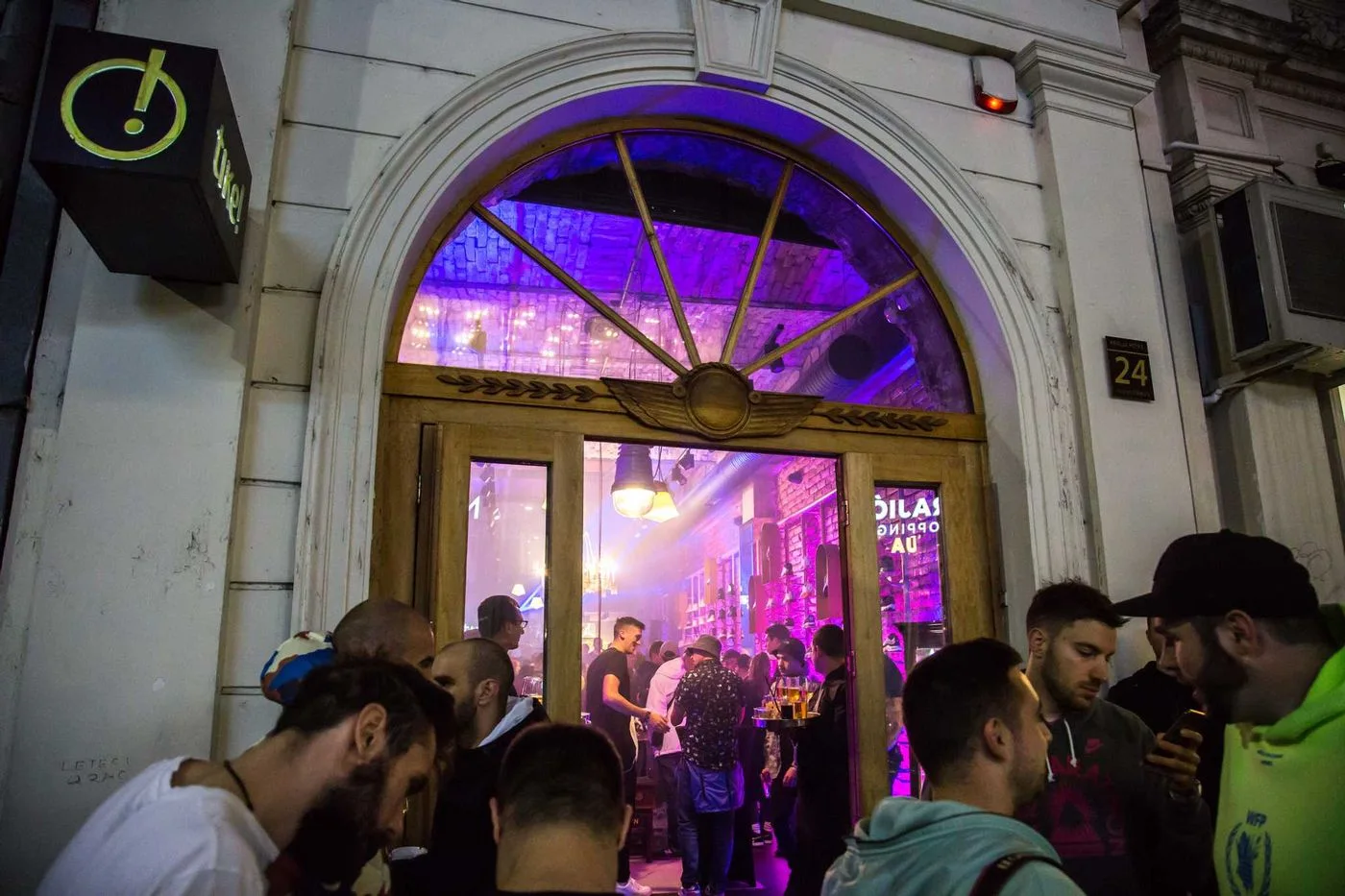People spilling out of the store's arched doorway onto the street at night, the interior lit purple and pink behind them
