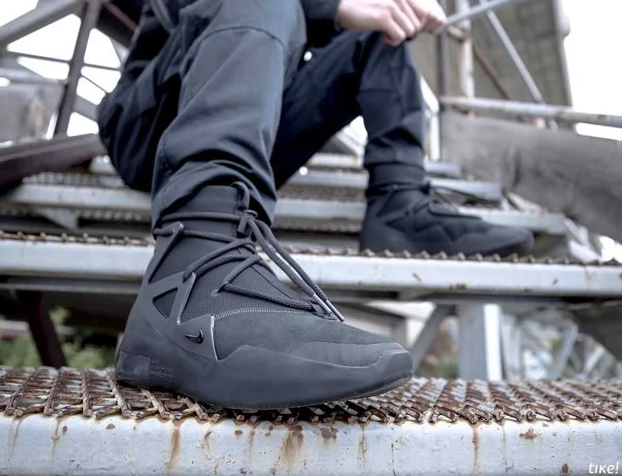 Close-up of the Nike Air Fear of God 1 Triple Black on a rusted metal walkway, the wrap-around lacing and matte suede upper filling the frame