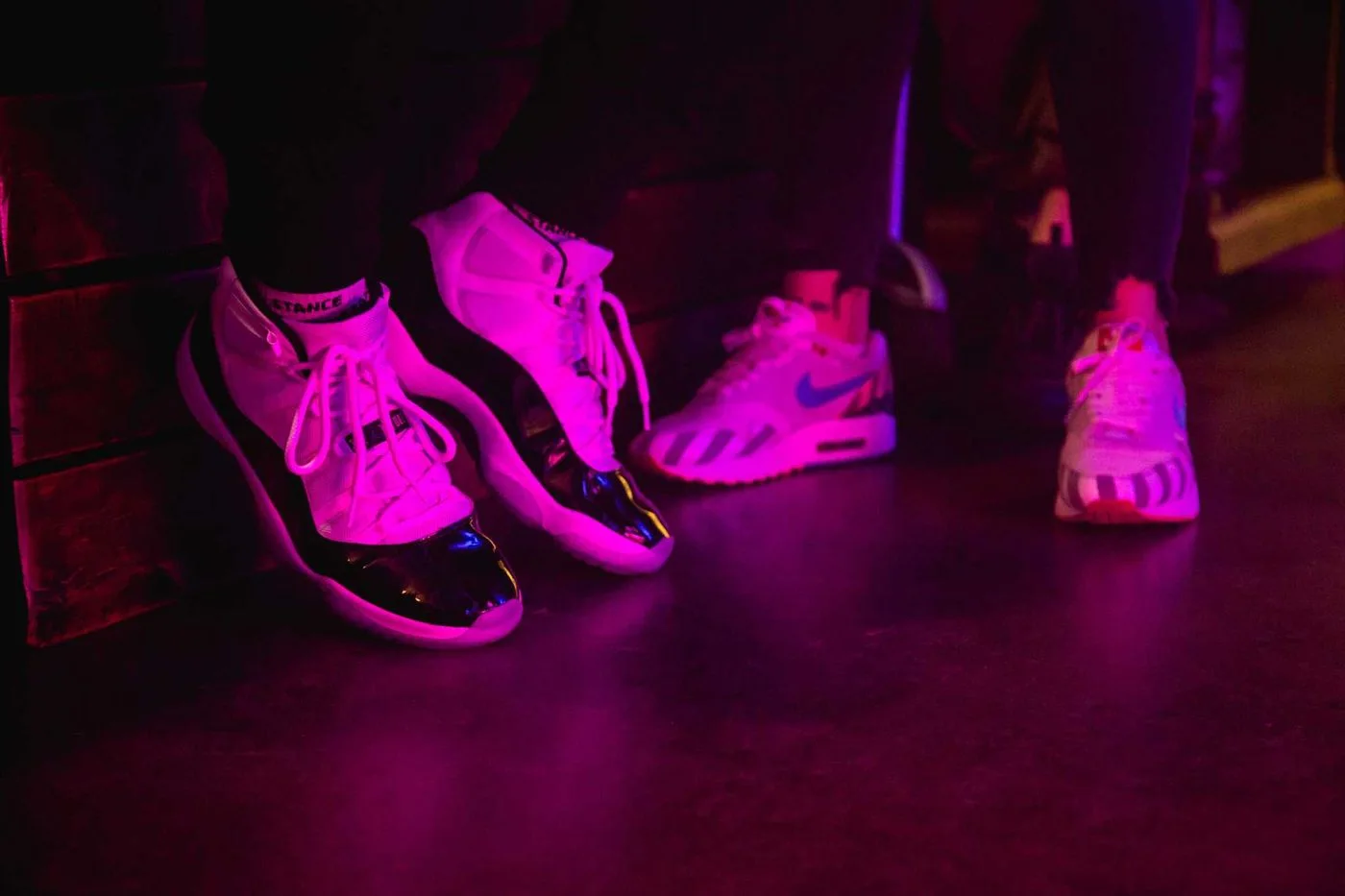 Close-up of sneakers on the floor under pink light, one pair of patent-leather high-tops in the foreground