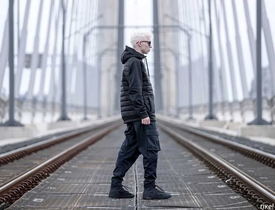 A model in an all-black hooded jacket and cargo trousers standing between the tram rails on a steel bridge, the truss receding into fog behind him