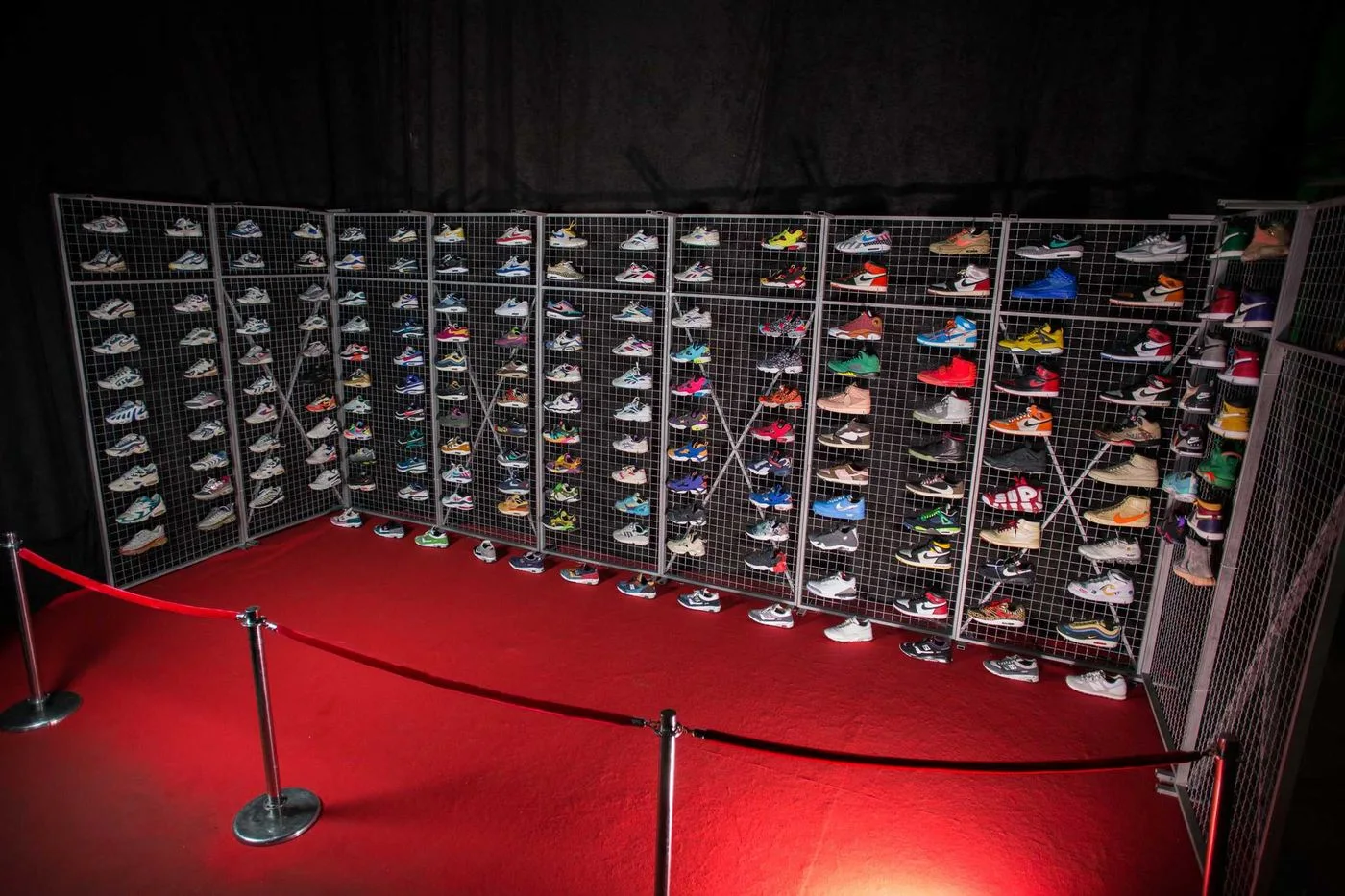 Hundreds of sneakers mounted on wire-mesh display panels in a darkened hall, a red rope barrier strung across the floor in front of them